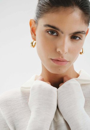 Mujer con cabello oscuro recogido, lleva pendientes de aro dorados y un top blanco de manga larga con textura, sosteniendo el cuello cerca de su garganta.