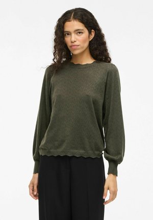 Femme aux cheveux bouclés portant un pull en maille à manches longues de couleur vert olive avec des bords festonnés et un pantalon noir sur un fond uni.