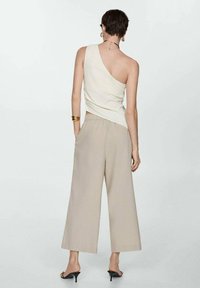 Žena s krátkými vlasy, oblečená v krémovém topu na jedno rameno, béžových širokých kalhotách, černých sandálech na podpatku a zlatých špercích, stojící zády.