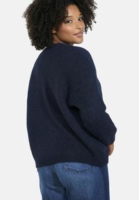 Tumši zila, pārāk liela, adīta džemperis ar teksturētu apdari, pazeminātiem pleciem un ribotām aprocēm, kombinēts ar gaiši zilām džinsām.