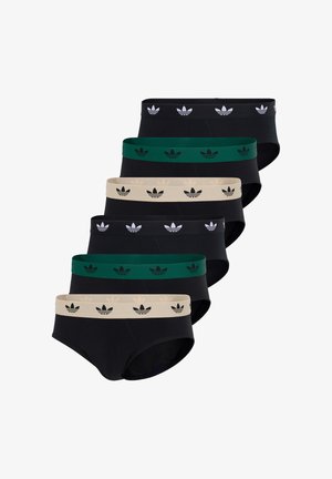 Młody mężczyzna o piegowatej skórze i dredach, ubrany w czarne slipy z zielonym pasem, stojący boso na białym tle.