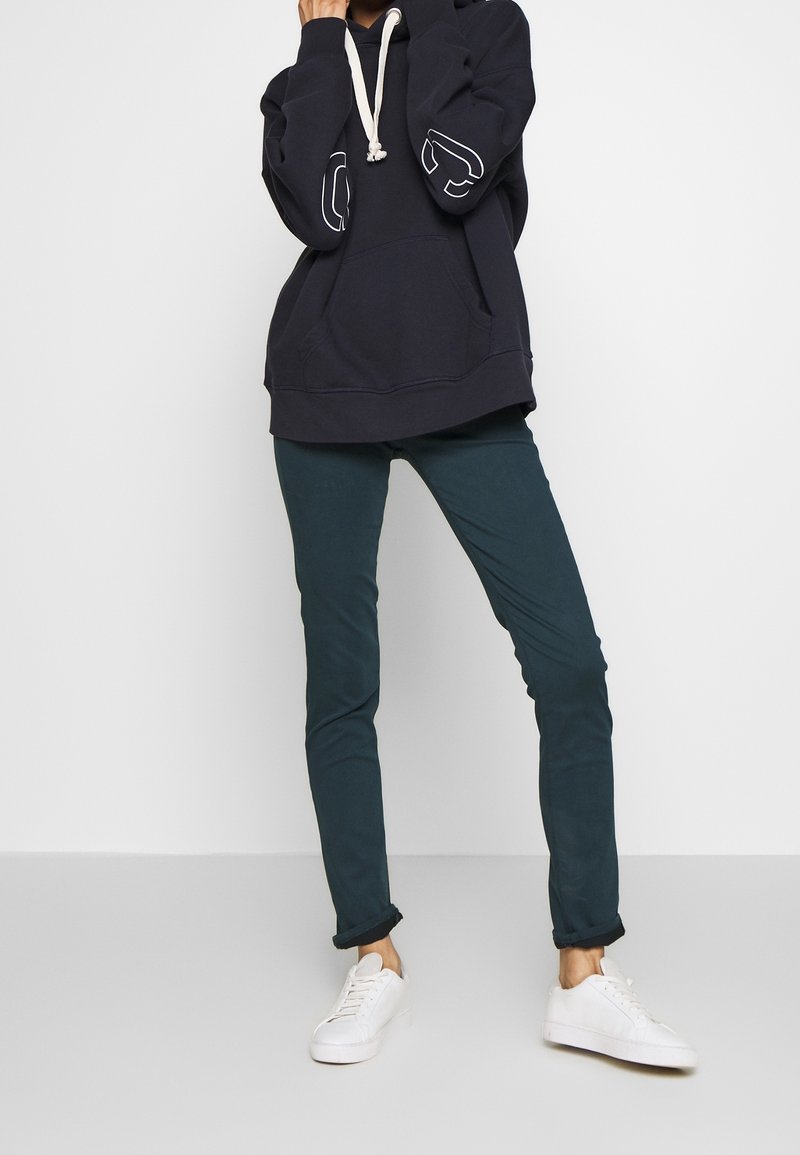 Marinblå hoodie med vita detaljer och dragsko; kombinerad med tealfärgade slim-fit byxor och vita sneakers. Avslappnad urban outfit visas.