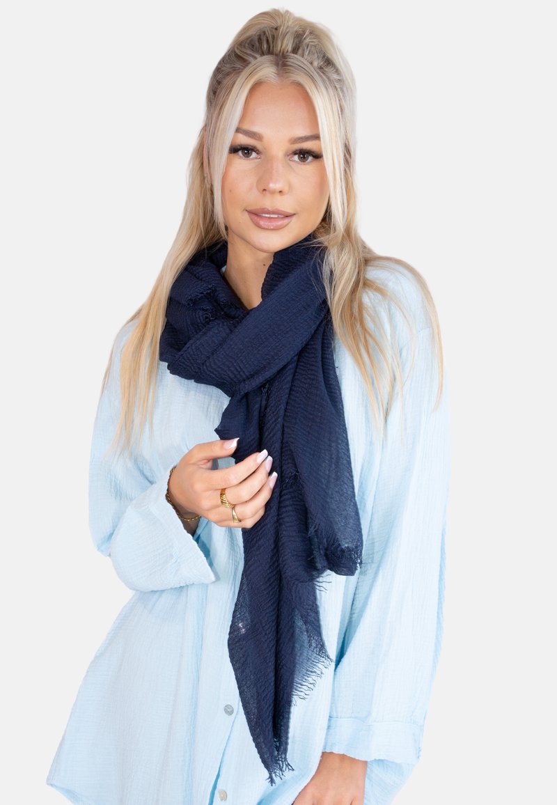 Écharpe tissée bleu marine avec une finition texturée, présentant des bords effilochés, portée lâchement autour du cou, associée à une chemise bleu clair.