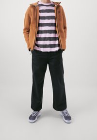 Veste marron zippée, t-shirt rayé rose et gris, pantalon noir et baskets grises avec semelles blanches. Tenue décontractée avec des textures variées.
