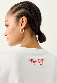 T-shirt bianca in cotone con testo ricamato rosso "Pop Off" sul retro. Il modello ha capelli intrecciati e orecchini a cerchio argentati.