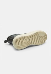 Zapatilla negra con parte superior texturizada, que presenta un diseño estampado y una suela de goma. La suela exterior es de color crema y tiene un patrón de dibujo que proporciona agarre.