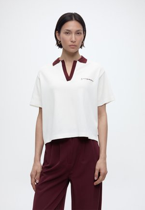 Γυναίκα με κοντά σκούρα μαλλιά που φοράει λευκό κοντομάνικο μπλουζάκι με μπορντό γιακά και αντίστοιχο μπορντό παντελόνι, στέκεται μπροστά σε απλό φόντο.