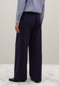 Marineblauwe wijde pijpen broek met een gladde textuur, voorzien van een achterzak en gecombineerd met een blauw-wit gestreept shirt. Zwarte schoenen maken de look af.