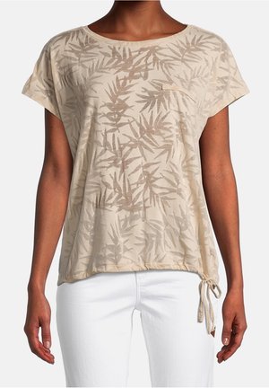 Beige kort shirt met bladpatroon, klein borstzakje en zijstrikzoom, gedragen met witte broek.