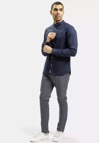 Granatowa koszula zapinana na guziki z kołnierzykiem stojącym, długimi rękawami i prostym krojem, zestawiona z jasnoszarymi zwężającymi się spodniami oraz białymi trampkami.