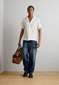 Korte mouwen gestippeld crèmekleurig shirt, vervaagde blauwe spijkerbroek, zwarte schoenen en een bruine leren sporttas in een minimalistische binnenomgeving.