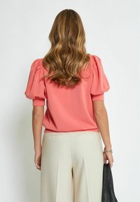 Top coral de manga abullonada con un corte holgado, combinado con pantalones de pierna ancha en un tono beige claro. Tejido texturizado, diseño minimalista y detalles sutiles.