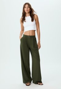 Top corto bianco e pantaloni larghi verde scuro con pieghe, dotati di passante per cintura e fibbia metallica, abbinati a sandali neri.