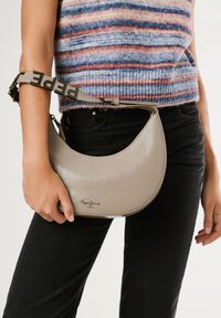 Osoba u crnim hlačama i prugastom džemperu drži bež torbu u obliku polumjeseca marki Pepe Jeans sa brendiranim remenom.