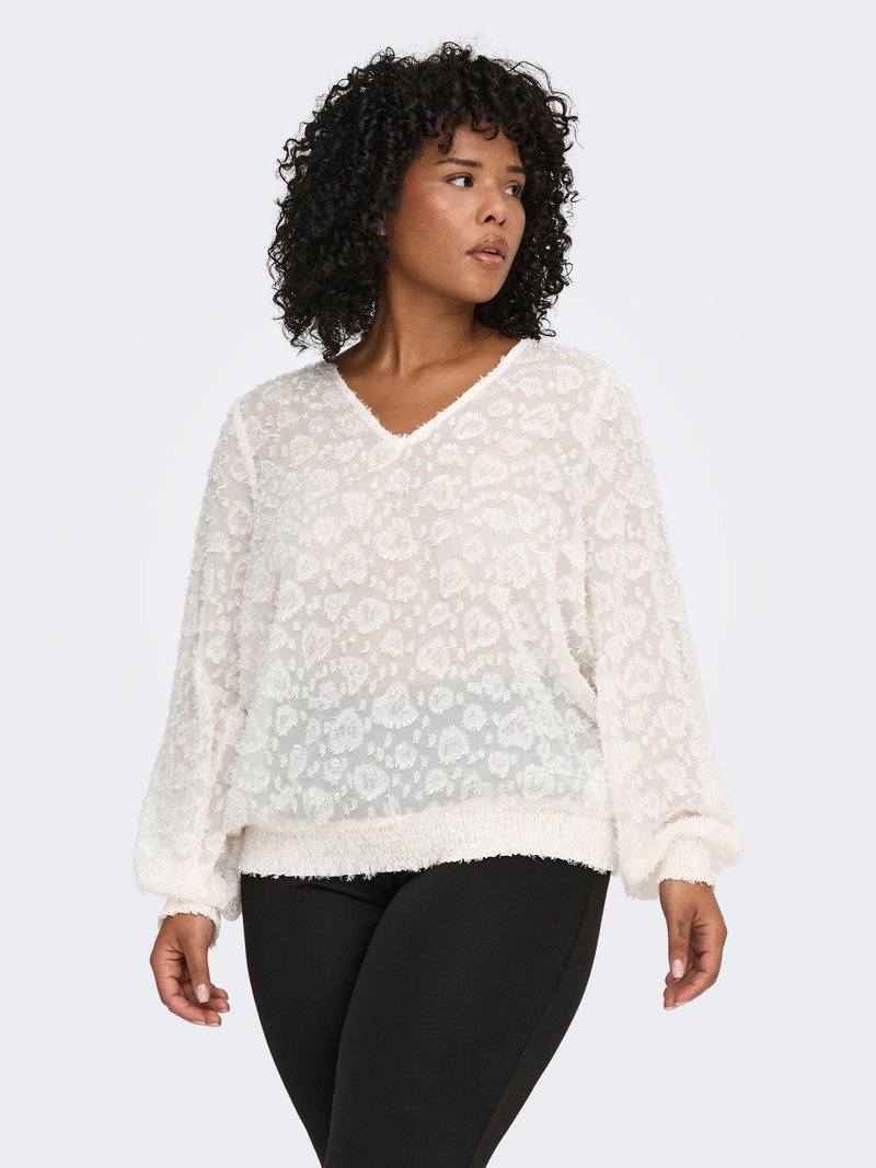 Vit texturerad blus med V-ringning, med ett blommönster, långärmad och med en fransig fåll. Bärs över svarta byxor.