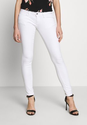 Vrouw draagt witte skinny jeans, zwart bloemenblouse en zwarte sandalen met hoge hakken, staand tegen een effen achtergrond.