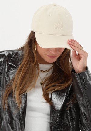 Beige baseball cap with embroidered logo, cotton material, curved visor, and ventilation holes, worn with a black shiny jacket.
