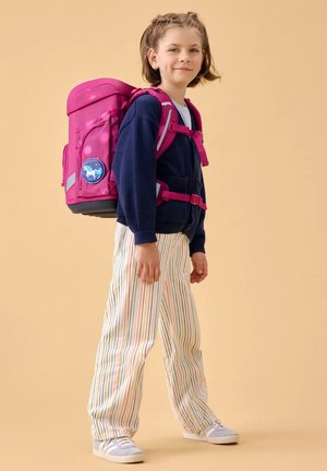Kind trägt marineblaue Strickjacke und gestreifte Hose, trägt einen großen rosa Rucksack mit einem Einhorn-Aufnäher und steht vor einem beigen Hintergrund.