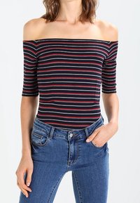 Bodysuit de hombros descubiertos y mangas cortas en color marino con rayas horizontales rojas y grises, combinado con unos vaqueros de tiro alto azul.