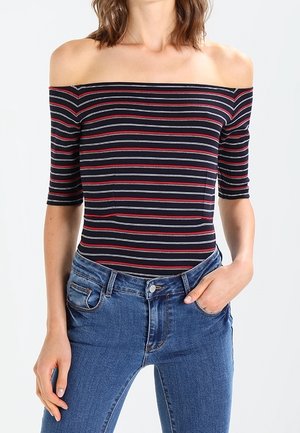 Off-shoulder, kortmouwen bodysuit in marineblauw met rode en grijze horizontale strepen, gecombineerd met high-waisted blauwe jeans.