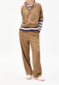 Donna che indossa pantaloni morbidi color cammello e un top abbinato sopra una maglietta a maniche lunghe blu e bianca a righe, con sneakers bianche.