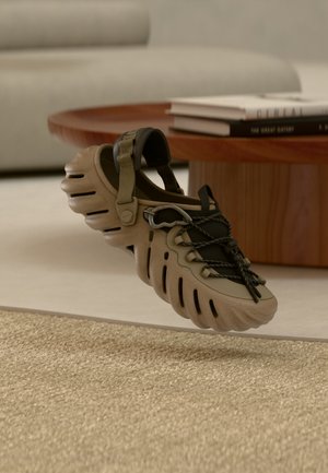 Bézs és fekete, nyitott hátú szandál vastag, texturált talppal, állítható pánttal és fekete fűzővel, faasztal mellett, könyvekkel megpakolva.