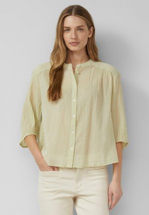Femme aux longs cheveux châtain clair portant un chemisier jaune clair boutonné à manches trois-quarts et un pantalon crème, debout contre un mur uni.