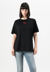 Young woman with straight dark hair wearing a black oversized t-shirt with a small red logo and light blue jeans, standing against white background.