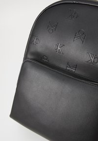Zaino in pelle nera con logo CK in rilievo, parte superiore arrotondata, tasca frontale con cerniera e texture liscia con finitura opaca.