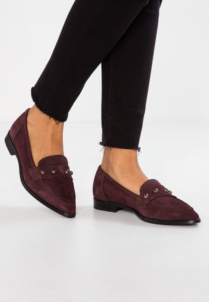 Personne portant un pantalon noir court et des mocassins en daim bordeaux avec des détails cloutés métalliques sur un sol blanc.