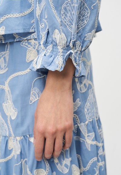 Hånd afslappet ved siden iført blå kjole med hvide broderede mønstre af muslinger og koraller, elastisk manchet med flæsedetalje.