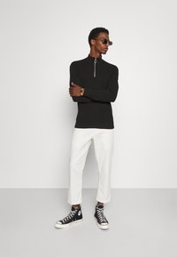 Svart langermet zip-up genser med et teksturert mønster, i kombinasjon med hvite cropped bukser og svarte høyhælte joggesko. Solbriller brukt som et aksent.