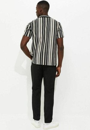 Mężczyzna stojący tyłem, ubrany w czarno-białą koszulę w krótkim rękawem z wzorem w zygzak, czarne spodnie i białe tenisówki na jednolitym tle.