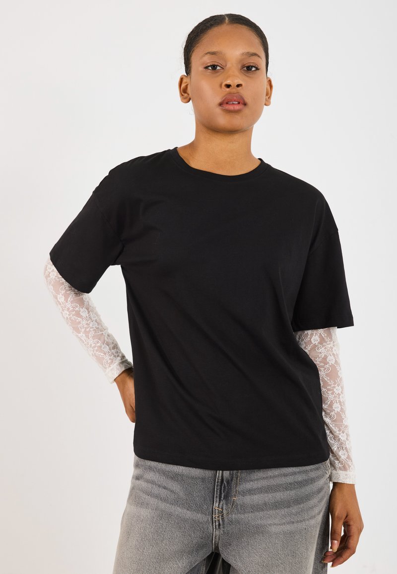 Svart bomull t-skjorte med korte ermer, lagd over en hvit blonde langermet topp. Grå denim jeans synlige ved livet. Enkelt design.