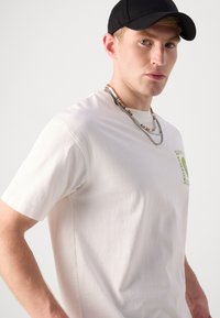 White cotton t-shirt featuring a green graphic print, short sleeves, and a relaxed fit, paired with layered necklaces and a black cap.