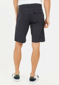 Marineblaue Shorts aus leichtem Stoff mit gerader Passform, mit Gesäßtaschen und genähtem Saum. Dazugehörig weiße Schuhe und grüne Akzente.
