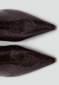 Zapatos de punta afilada en imitación cuero marrón oscuro con textura de cocodrilo grabada, presentando costuras visibles a lo largo de las uniones.