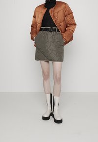 Veste orange gonflée, col roulé noir, jupe matelassée olive avec une ceinture noire, et bottes blanches montantes avec des accents noirs.