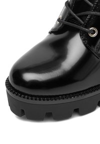 Černé lakované kožené kotníkové boty s zaoblenou špičkou, texturovanou podrážkou a kulatými šněrovadly procházejícími stříbrnými očky. Mají vroubkovaný okraj.