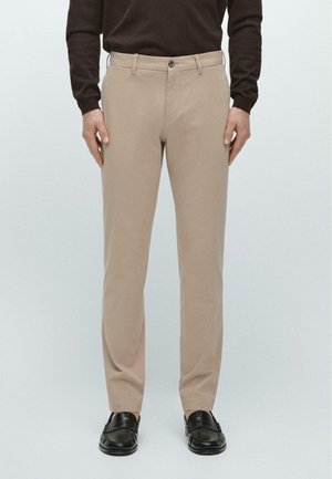 Mężczyzna ubrany w beżowe, dopasowane spodnie, czarne mokasyny oraz ciemnobrązową bluzkę z długim rękawem, stojący na jednolitym tle.