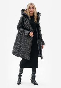 Zwarte, waterdichte pufferjas met grote capuchon, diamantpatroon textuur en zijzakken, gecombineerd met een lange zwarte jurk en kniehoge laarzen.