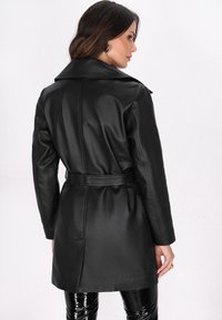 Zwarte leren trenchcoat met een riem in de taille, reverskraag en lange mouwen. Glad oppervlak, knielang en passend model.