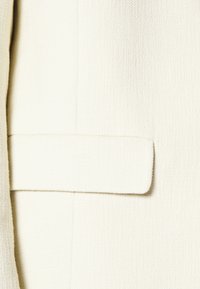 Gros plan d'une veste en tissu blanc texturé avec un détail de poche à rabat horizontal à l'avant.