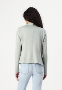 Svetlo zelena uska blazer jakna z gladko teksturo, dolgimi rokavi in ovratnikom s poklopcem. Nosi se čez svetlo modre denim kavbojke.