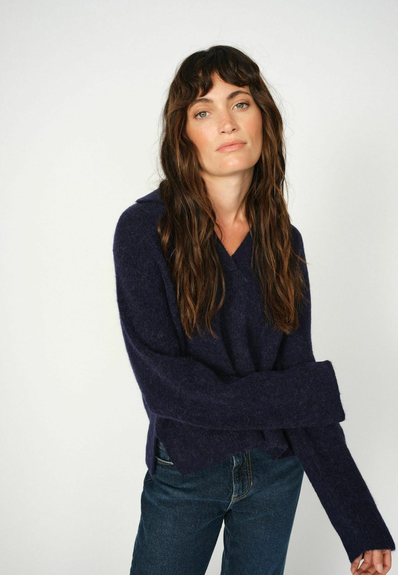 Donkerblauwe, losgebreide trui met een capuchon en brede mouwen, gemaakt van zachte, gestructureerde stof. Draag met strakke donkergekleurde jeans.