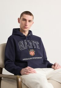Sudadera azul marino de algodón, con texto blanco "GANT", un parche de logo y un bolsillo delantero. Se lleva con pantalones de color claro.