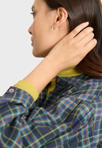 Camicia in tartan navy con dettagli verdi, dotata di collo a camicia, chiusura con bottoni e maniche lunghe, indossata da una persona che si tocca il collo.