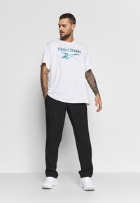 T-shirt branca de algodão com o logo da Reebok em turquesa, combinada com calças pretas e sapatilhas brancas; o modelo tem tatuagens e cabelo curto.