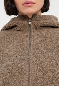 Καφέ φλις μπουφάν με υφή, αφράτη επιφάνεια, που διαθέτει φερμουάρ μπροστά και κουκούλα. Το υλικό είναι μαλακό και άνετο.