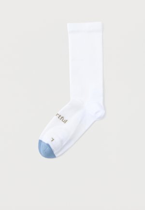 Balta komandas zeķe ar gaiši zilu purngala daļu un zelta krāsas tekstu pie purngala, uz gaiši pelēka fona.